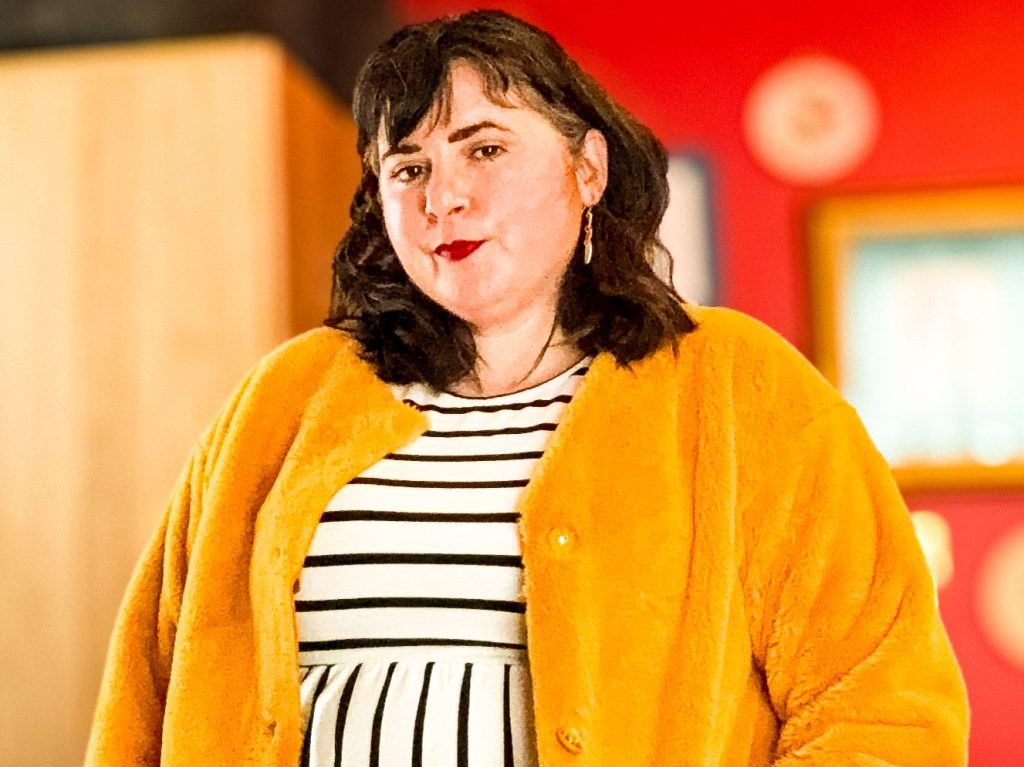 A brown-haired woman wearing a yellow faux fur coat and a black-and-white striped dress. 