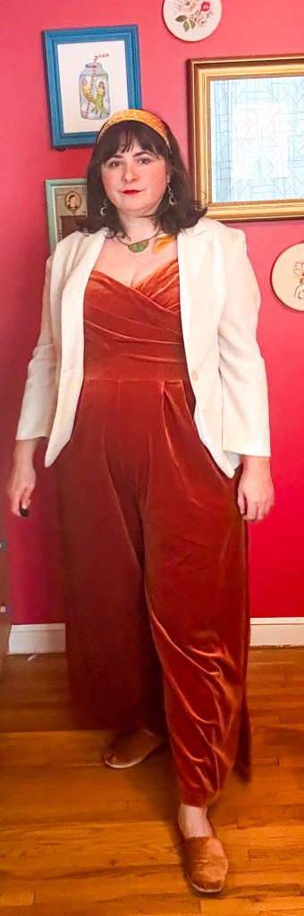 A 1970s-inspired outfit featuring a orange velour jumpsuit, white linen blazer and an orange silk headband. 